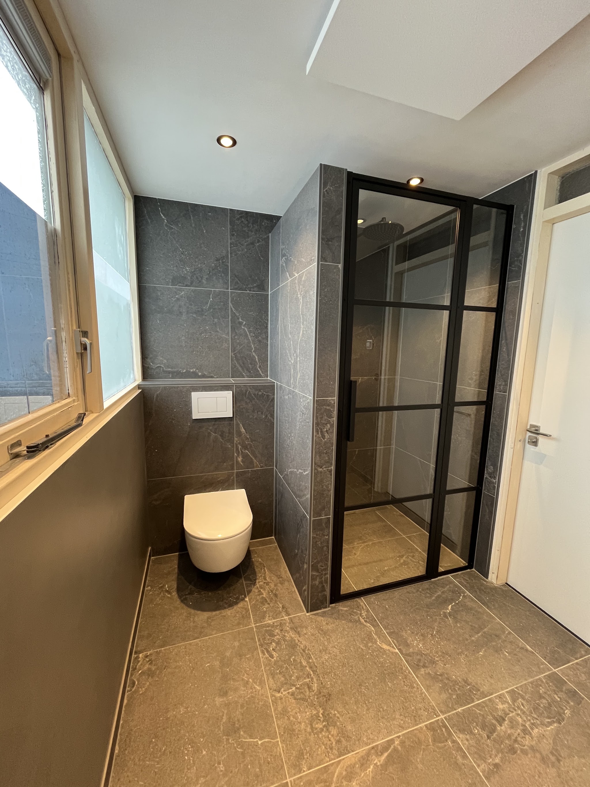 Opgeleverde badkamer met antracietkleurig marmerlook tegelwerk, hangend toilet en een zwart omlijste glazen douchewand