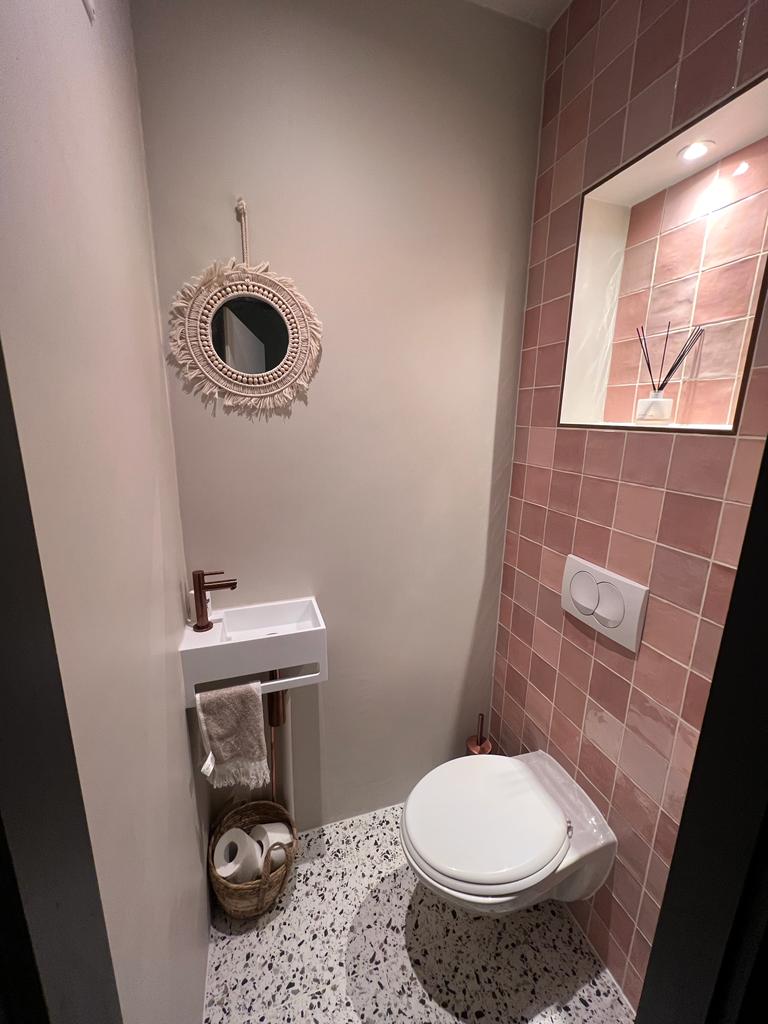 Toilet met terracotta tegels, terrazzovloer, fonteintje met koperen kraan en een ronde spiegel
