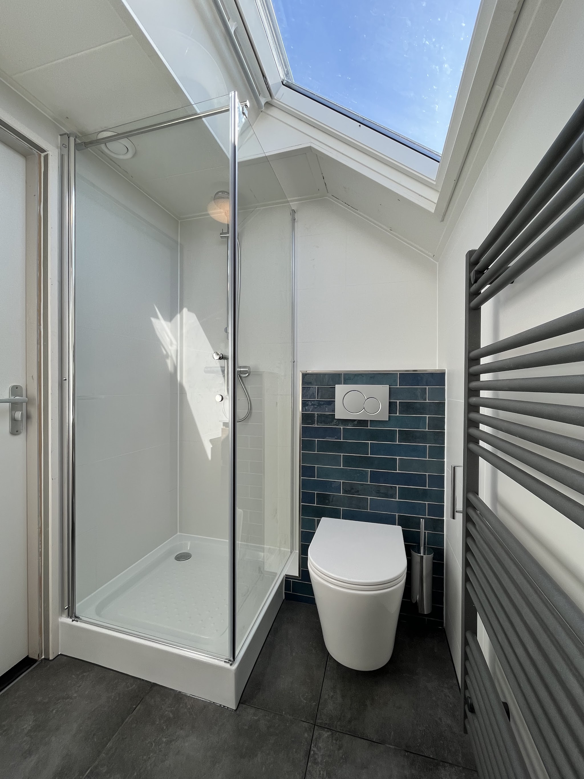 Badkamer onder een schuin dak met douchecabine, hangend toilet tegen diepblauwe tegels en een dakraam