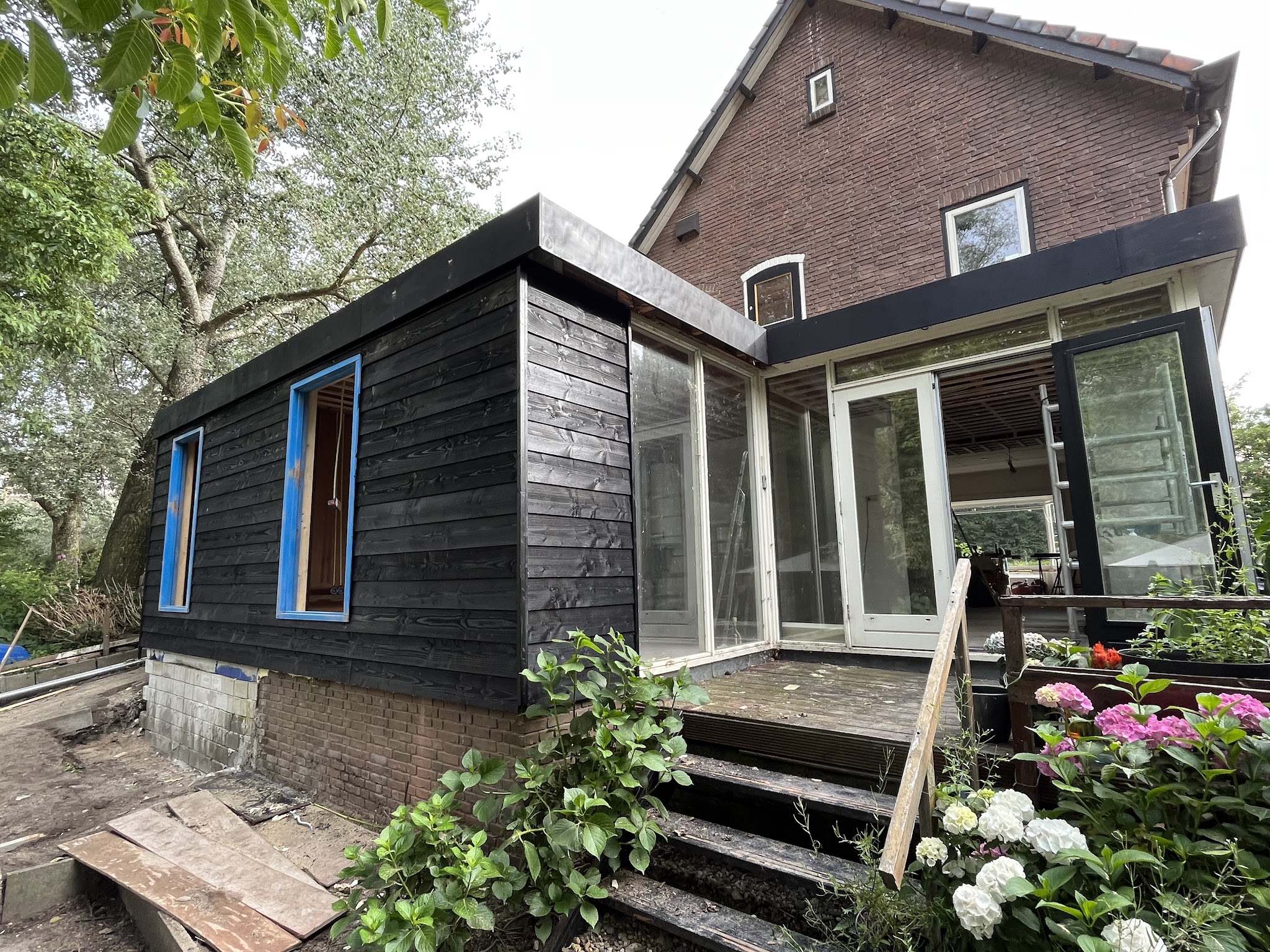 Aanbouw in aanbouw met zwart gebeitste houten gevelbekleding en nieuwe glazen schuifpui tegen een woning